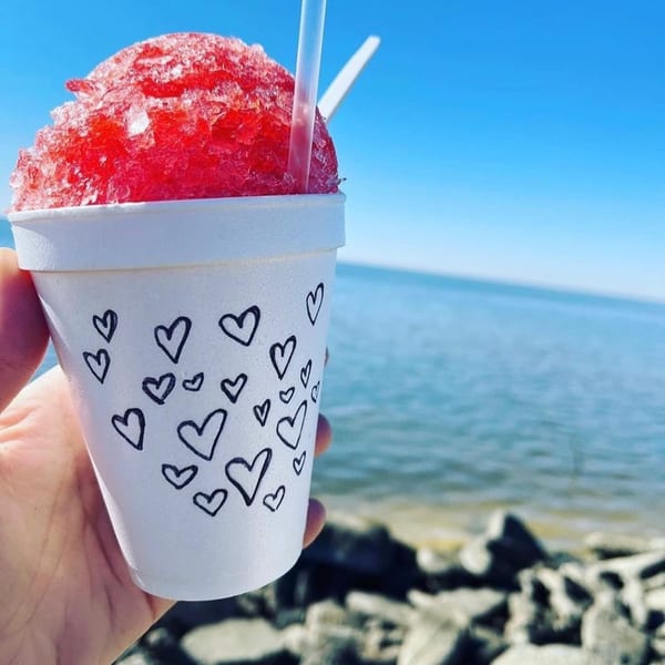 Hand holding a white cup with black hearts, filled with red shaved ice, by the ocean.