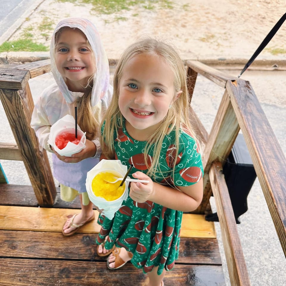 Two smiling young girls holding colorful snow cones on a wooden deck.