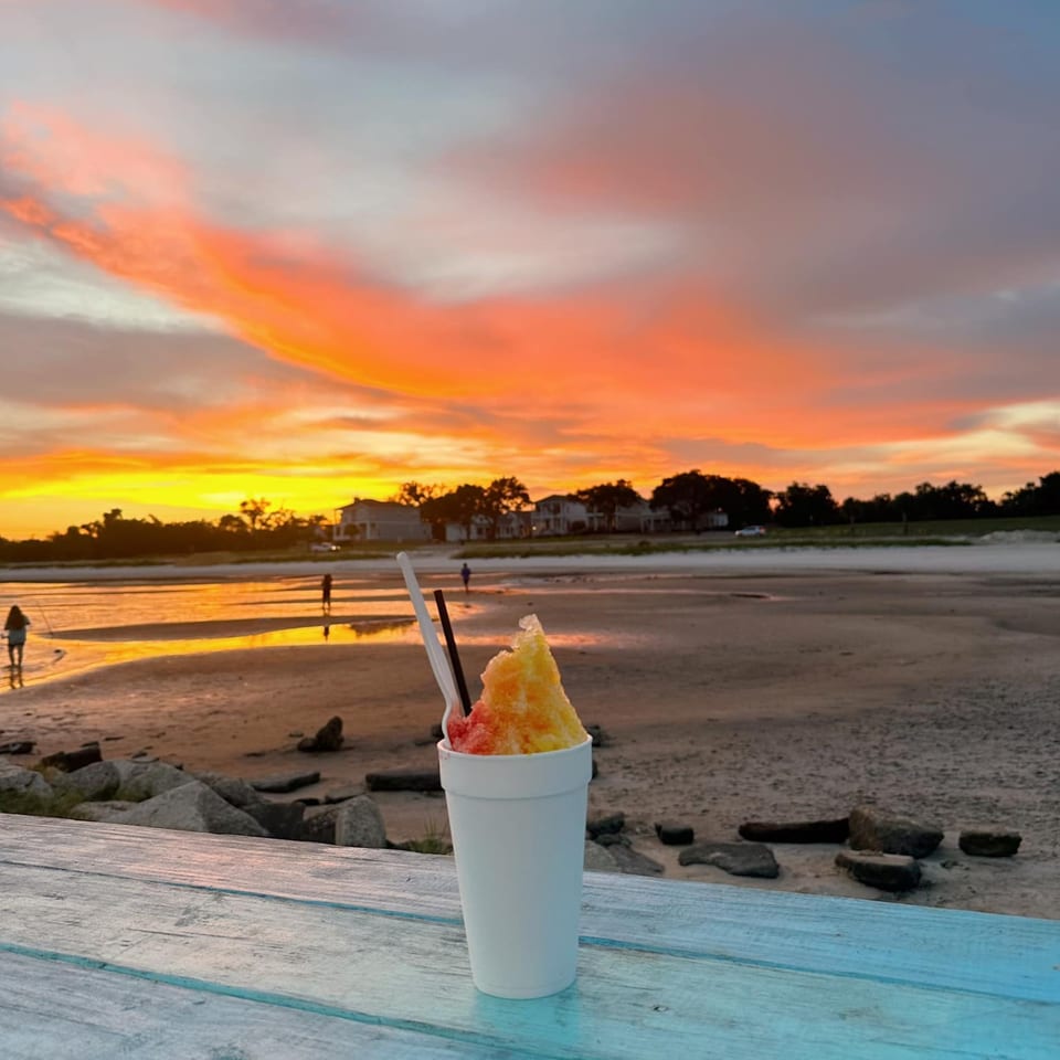 Shaved ice on a blue table with a vibrant orange sunset over a beach and distant houses.