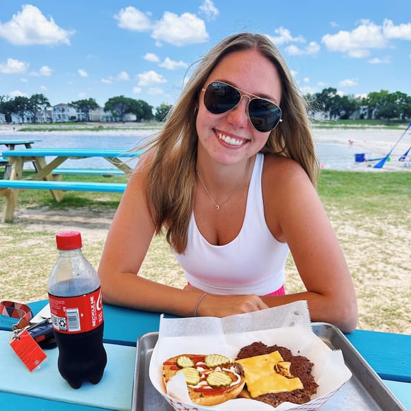 A smiling woman in sunglasses sits at a blue picnic table with a burger and soda by the water.