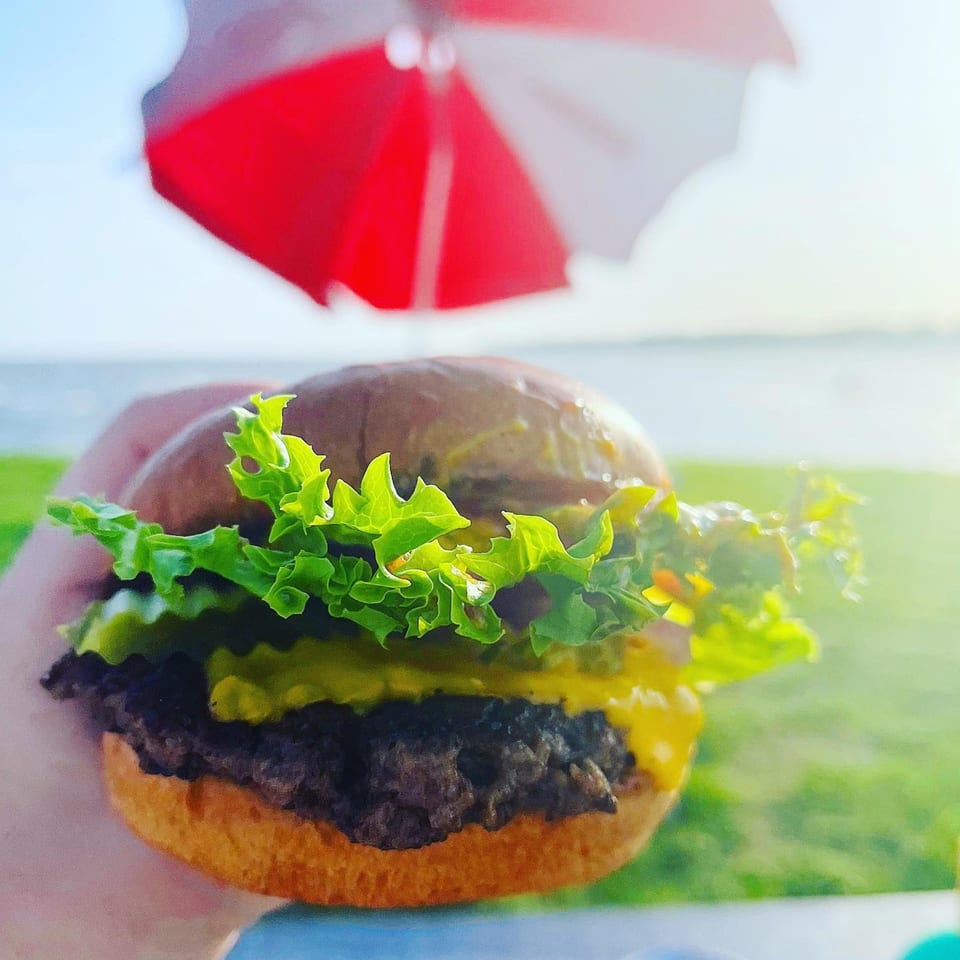 A Sea Level burger with lettuce and cheese, held outdoors with a red umbrella and water in the background.