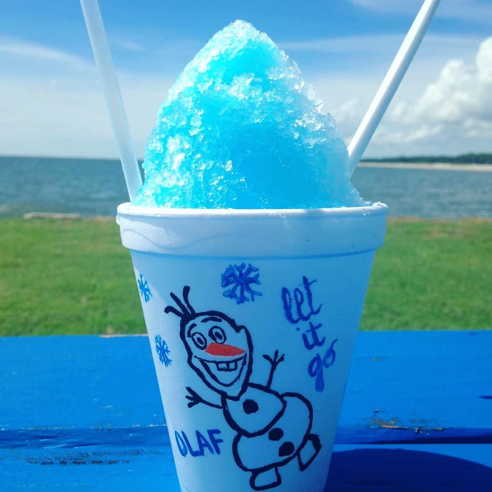 Blue shaved ice in an Olaf cup on a blue table, with a lake and sky in the background.