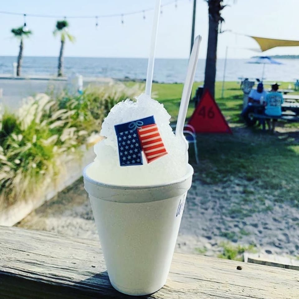 Shaved ice with an American flag pick and two spoons on a wooden railing overlooking the ocean.