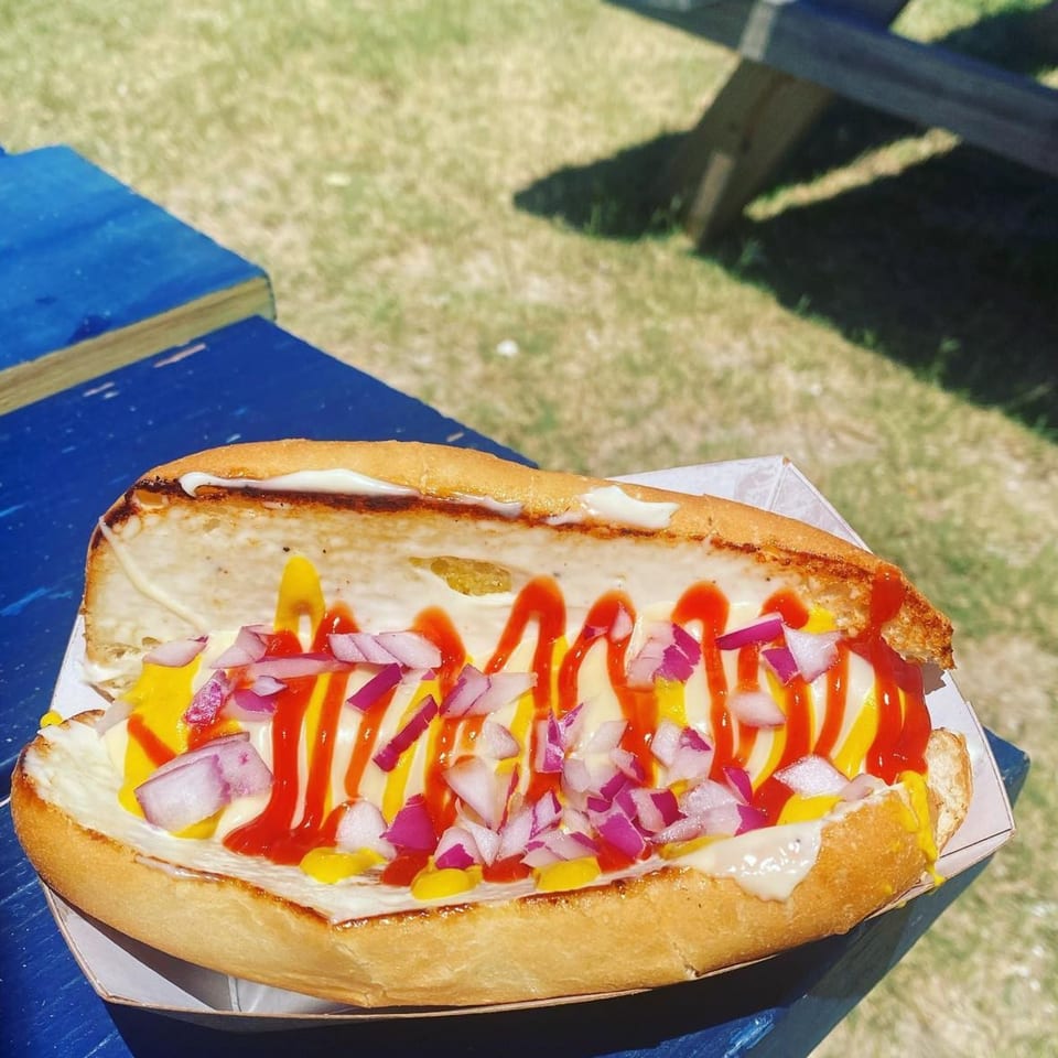 Hot dog with mayonnaise, mustard, ketchup, and diced red onions on a blue picnic table.