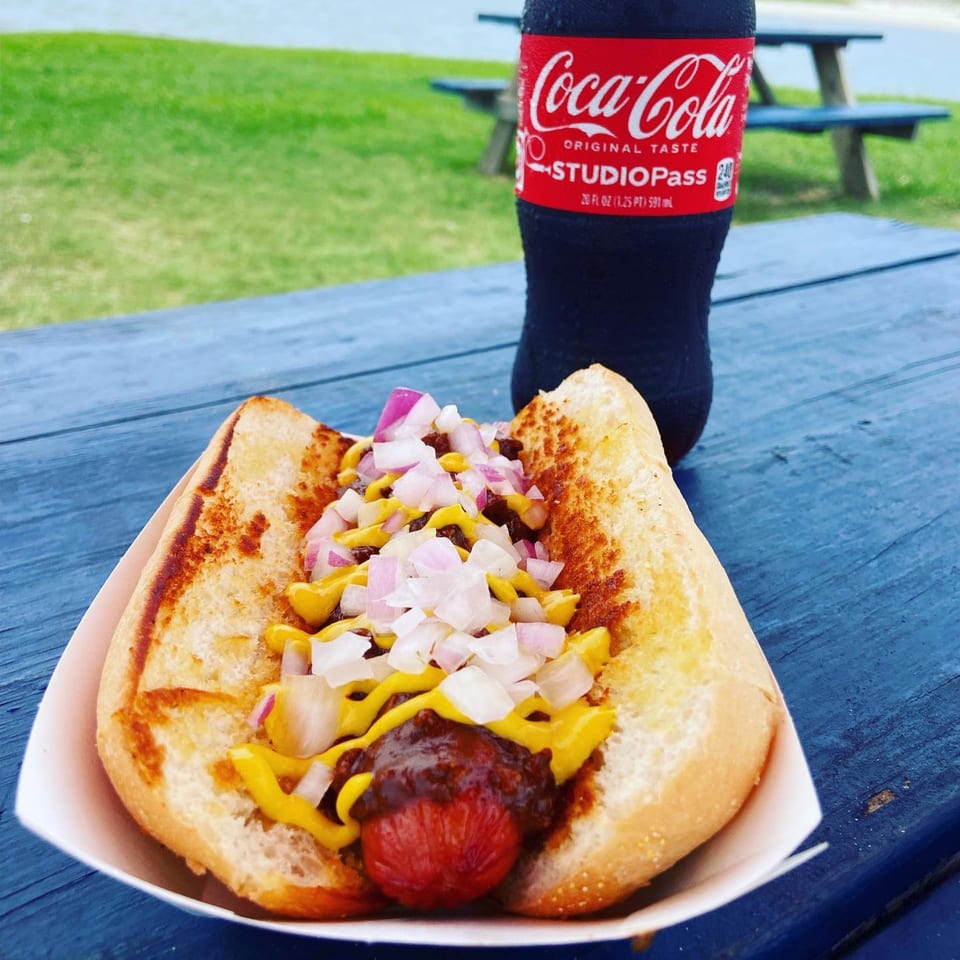 A chili cheese hot dog with diced onions and mustard, next to a Coca-Cola bottle on a blue picnic table.