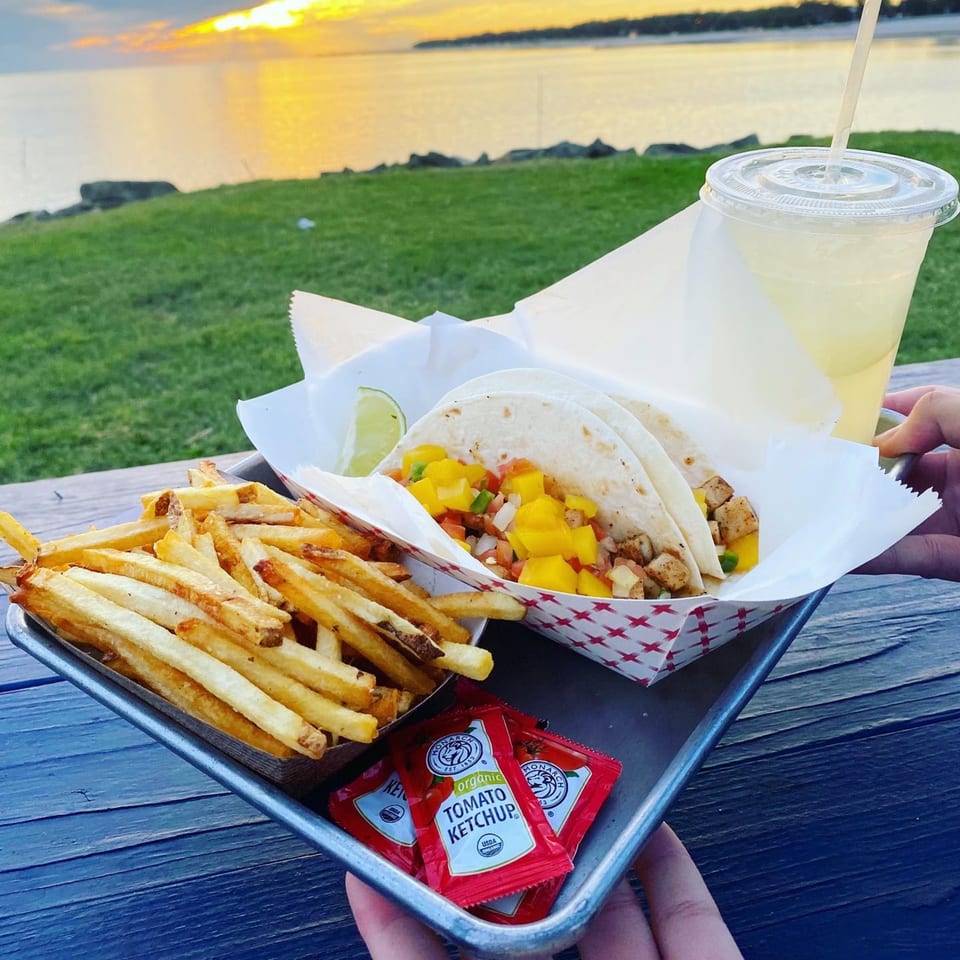 Fish tacos with mango salsa, fries, and a drink on a tray overlooking a sunset over water.