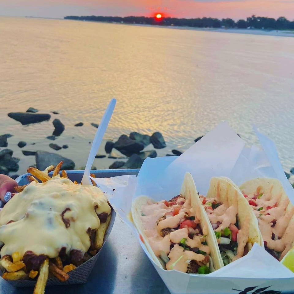 Chili cheese fries and tacos on a tray with a sunset over the water in the background.