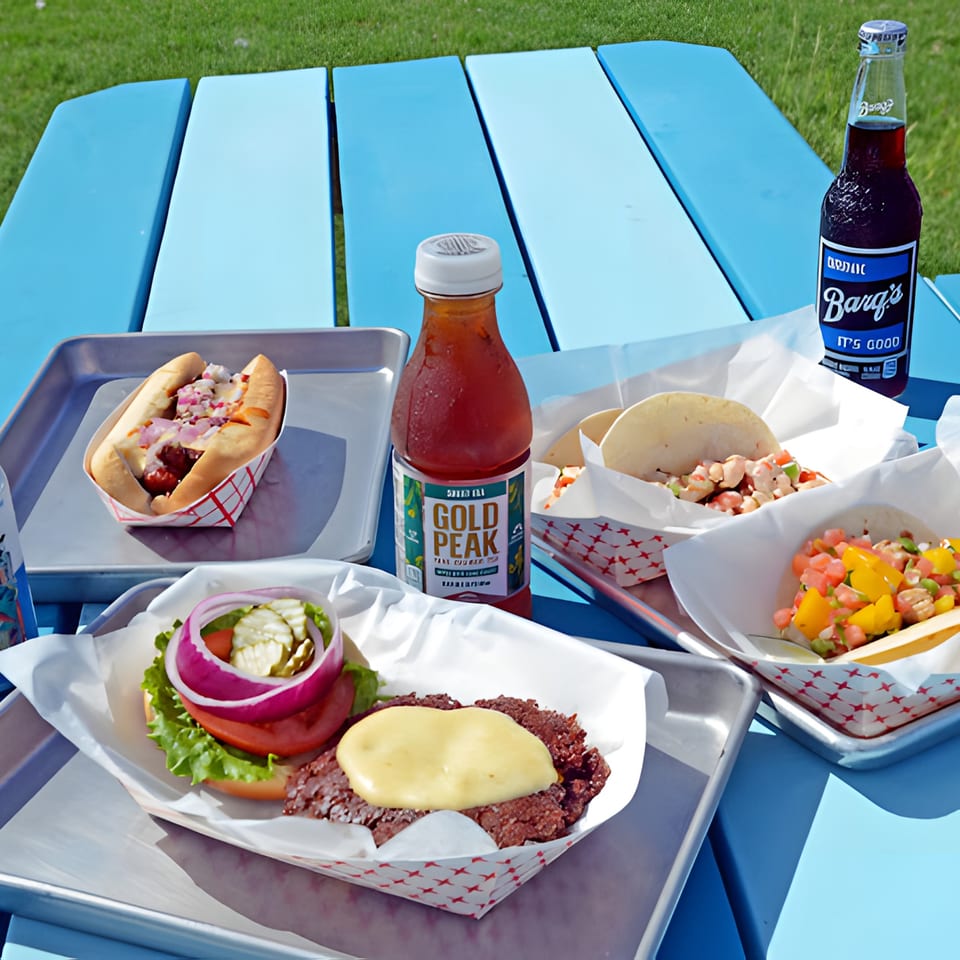 A burger, hot dog, tacos, and drinks on a blue picnic table with green grass in the background.